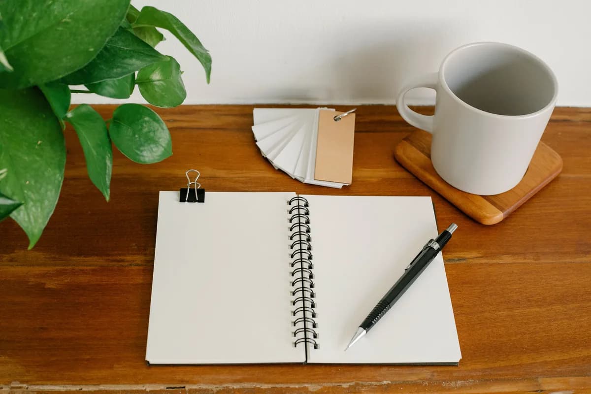 Open notebook and pen on a wooden desk beside a mug and a plant