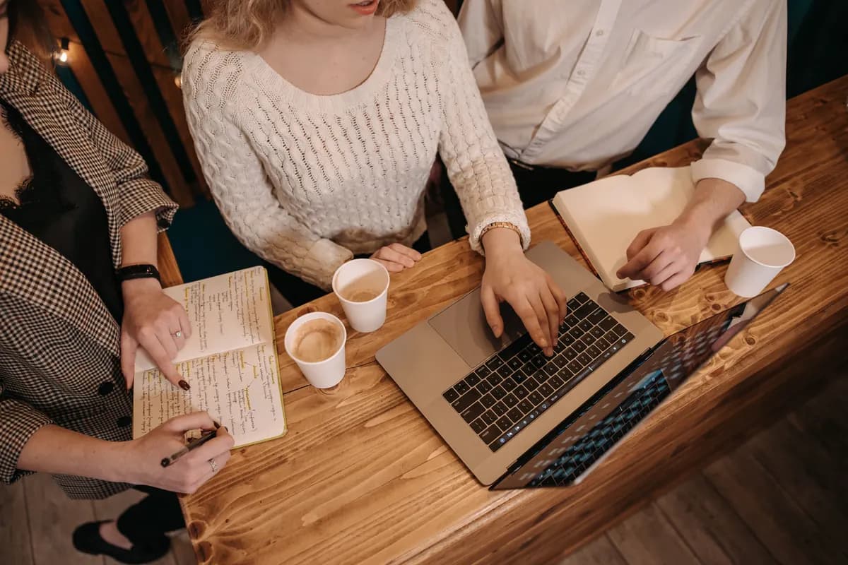 Colleagues' hands over notebooks, coffee and a laptop at a wooden table as they review material together